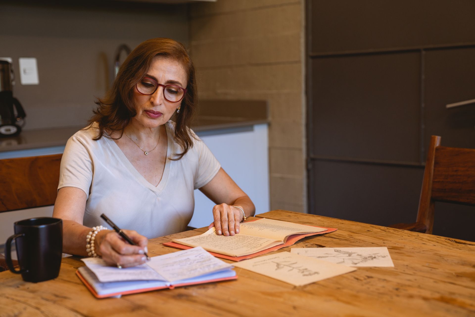 Una donna adulta scrive appunti al tavolo di cucina, fra i libri e una tazza di caffè