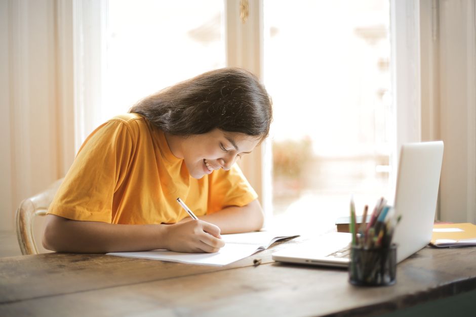 Una ragazza sorride mentre studia a casa con il portatile e un quaderno di appunti