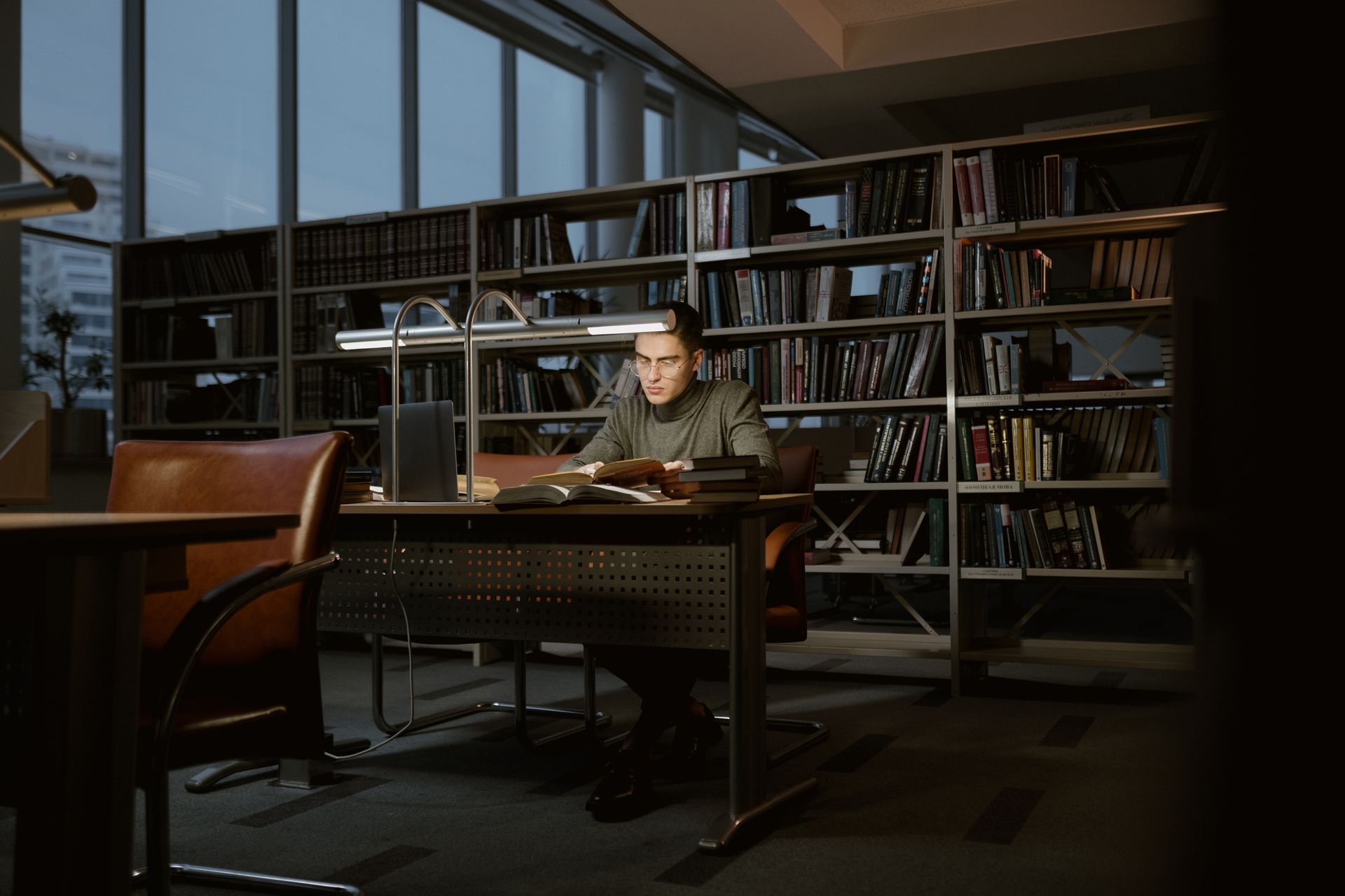Un uomo studia concentrato fra i libri di una biblioteca, nella luce calda di una lampada